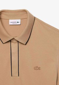 Beżowa koszulka polo z teksturowanej tkaniny, z czarnym dekoltem i dwoma guzikami. Logo Lacoste jest wyhaftowane na lewej piersi.