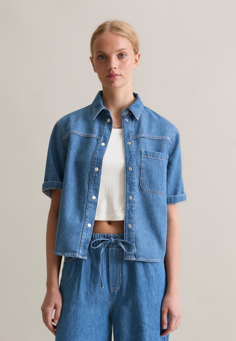 Denim shirt in light blue, short sleeves, button-up front, single chest pocket, worn over a white ribbed crop top, paired with matching denim bottoms.