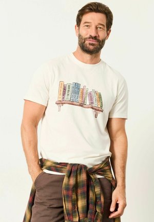 Mann trägt weißes T-Shirt mit buntem Bücherregal-Aufdruck, braune Hose und kariertes Hemd um die Taille gebunden, steht vor hellem Hintergrund.
