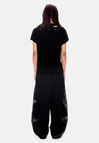 Άτομο με μακριές κοτσίδες στέκεται γυρισμένο προς τα πίσω, φοράει μαύρο φθαρμένο μπλουζάκι και φαρδιά μαύρη παντελόνια με λευκά σχέδια.