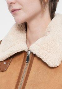 Hnědá semišová bunda s krémovým kožešinovým límcem, stříbrným zipem a knoflíkovými detaily, s hladkou texturou a strukturovaným designem.