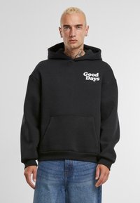 Zwarte fleece hoodie met de tekst "Good Days" in het wit op de borst, een buideltas, en een capuchon met trekkoord. Draagt met lichtblauwe jeans.