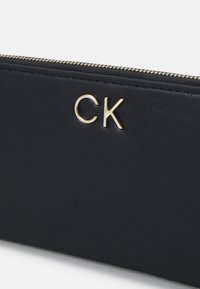 Portafoglio in pelle blu navy con chiusura a zip e logo dorato "CK". Texture liscia senza motivi visibili o accessori aggiuntivi.