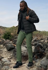 Chaqueta inflable negra sobre una camiseta negra, pantalones cargo verdes y zapatillas negras. Entorno al aire libre con terreno rocoso y vegetación al fondo.