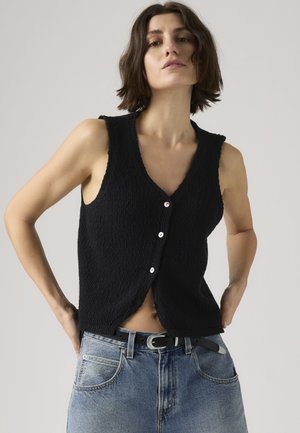 Mujer con cabello corto y castaño lleva un chaleco negro sin mangas abotonado y jeans azul claro con cinturón negro, posando con las manos en las caderas.