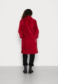 Kiharatukkainen henkilö, joka pukeutunut punaiseksi keskipitkän tekoturkistakin, mustiin housuihin ja mustiin saappaisiin, seisoo kasvot valkoista seinää kohti.