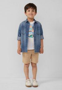 Kind dat een lichtblauw denim overhemd draagt over een witte T-shirt met opdruk, beige korte broek en witte schoenen met bruine zolen. Eenvoudige en casual outfit.