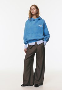 Felpa oversize blu con tasca frontale e testo grafico, indossata sopra una camicia a righe blu chiaro, abbinata a pantaloni larghi marroni.