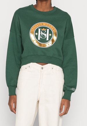 Osoba nosząca zieloną krótko skrojoną bluzę z kapturem z złotym okrągłym logo z napisem "Hollister Southern California" oraz białe spodnie z wysokim stanem.
