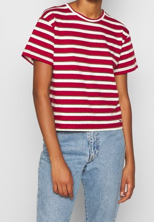 Persoon draagt een rood-wit horizontaal gestreept T-shirt met korte mouwen en lichtblauwe hoog getailleerde jeans tegen een effen achtergrond.