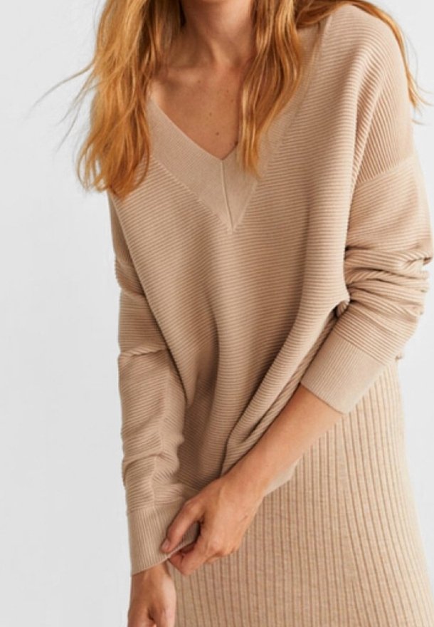 Femme portant un pull beige côtelé à col en V et une jupe côtelée assortie, ajustant la manche avec sa main droite.