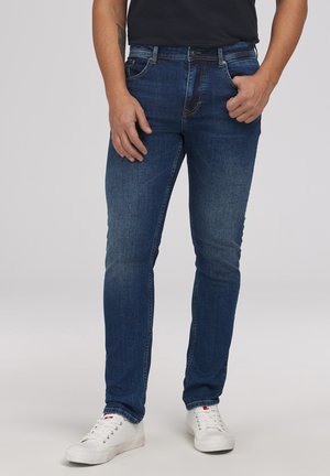 Mężczyzna ubrany w dopasowane niebieskie dżinsy i białe trampki, stojący z jedną ręką w kieszeni na jednolitym tle.