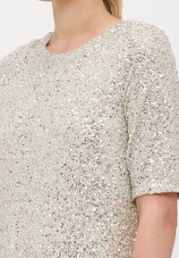 Kortärmad topp täckt av silver- och guldpaljetter; rundad halsringning; texturerad yta; glanseffekt; inga synliga hårdvaror eller mönster.