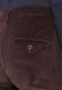 Gros plan sur la poche arrière d’un pantalon en velours côtelé marron avec fermeture par bouton, porté avec un haut sombre.