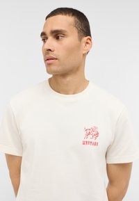 T-shirt en coton crème avec un col rond. Présente un graphique rouge d'un cheval et le texte "MUSTANG" sur la partie gauche de la poitrine. Manches courtes.