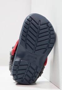 Szary kapcie z tkaniny z teksturowaną czarną gumową podeszwą z rowkami zapewniającymi przyczepność. Czerwony akcent po boku w pobliżu pięty.