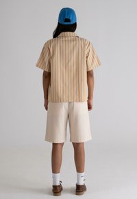 Short-sleeved, striped shirt in beige and orange, paired with beige shorts. Accessories include a blue cap and brown shoes.