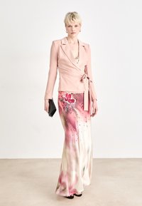 Rosa omslagsblazer med lange ermer, kombinert med et flytende skjørt med akvarellblomster mønster og paljetter; svart clutch holdt i hånden.