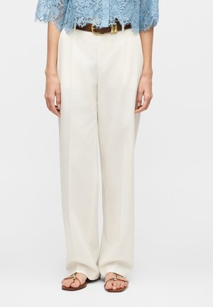 Frau trägt cremefarbene, weit geschnittene Hose mit hoher Taille und braunem Gürtel, blaues Spitzentop und beige Sandalen mit goldenen Akzenten.