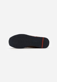 Brown sneaker with a textured upper, featuring a black rubber outsole and red accent detail. The tread pattern is wavy for traction.