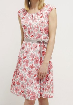 Femme portant une robe sans manches mi-genoux avec un motif floral rose et blanc et une ceinture grise nouée à la taille.