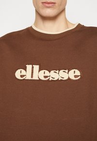 Hnědá bavlněná mikina se béžovým vyšitým logem „ellesse“ na přední straně, kulatý výstřih s kontrastním žebrovaným límcem.