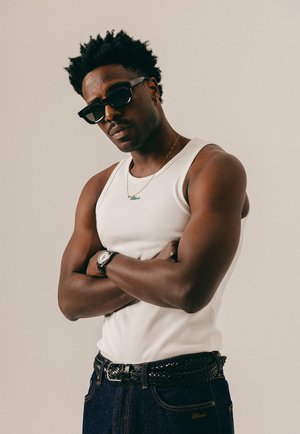 Mann med armene i kors, iført svarte solbriller, hvit singlet, gullkjede, klokke og mørke jeans mot en nøytral bakgrunn.