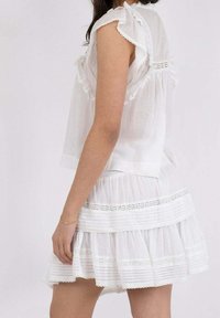 Donna che indossa una blusa bianca senza maniche con volant e una gonna a strati coordinata, in piedi di lato contro uno sfondo chiaro e uniforme.