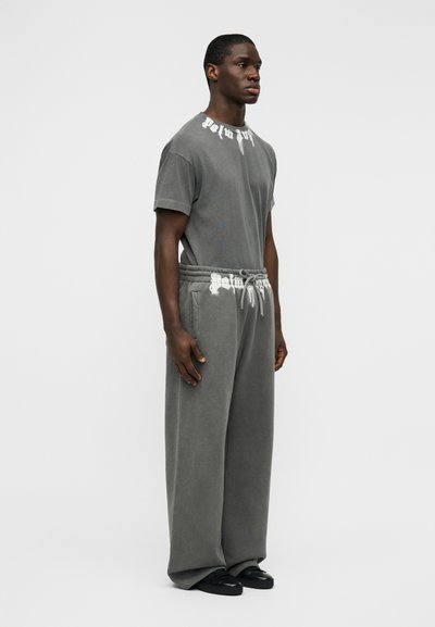 Γκρι βαμβακερό μπλουζάκι και αντιστοιχές φαρδιές φόρμες με αθλητική υφή. Το μπλουζάκι φέρει έντονα λευκά γράμματα· το παντελόνι έχει κορδονάκι στη μέση με ξεφτισμένες λεπτομέρειες.