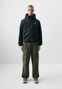 Casaco de fleece preto com fecho zip e capuz, combinado com calças cargo largas em verde-oliva e sapatilhas cinzentas com detalhes brancos.