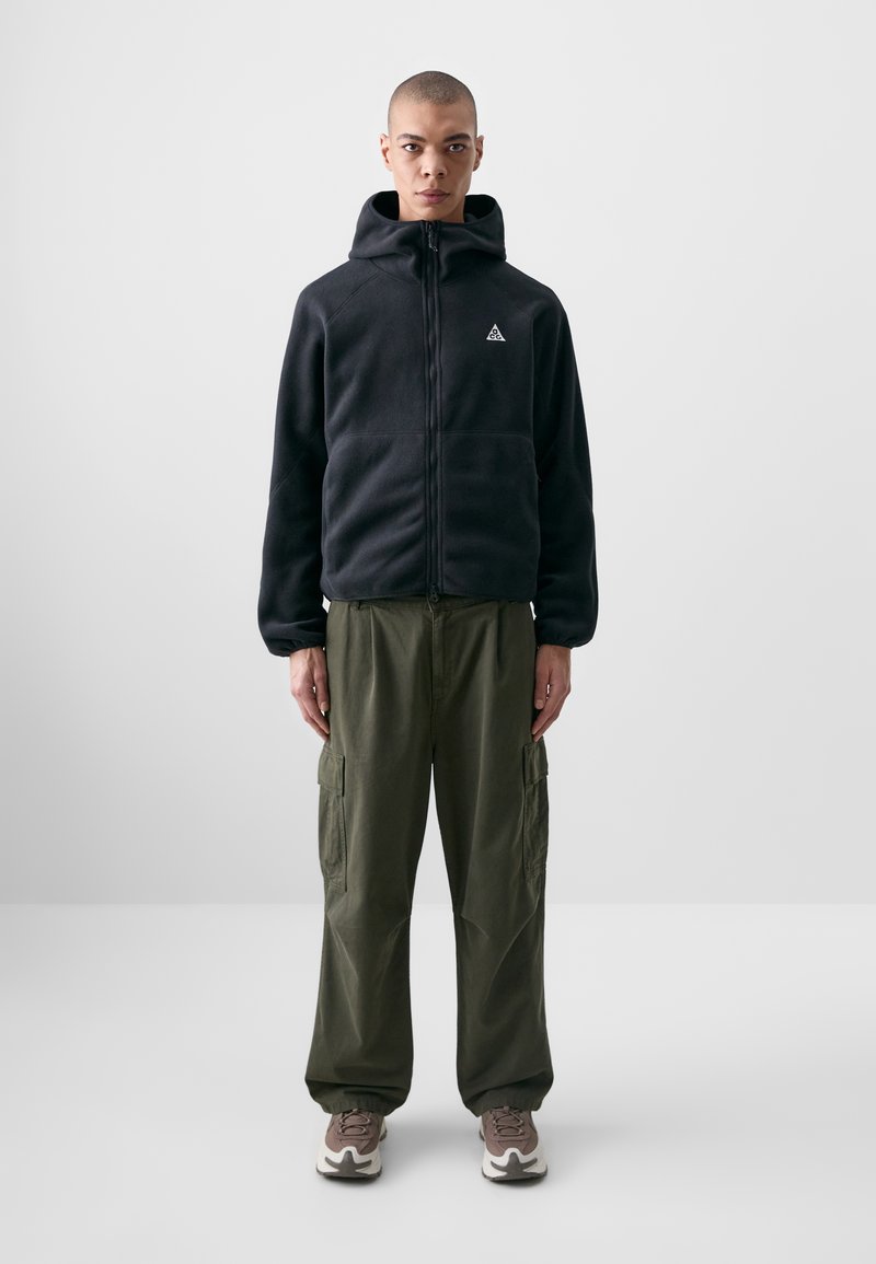 Casaco de fleece preto com fecho zip e capuz, combinado com calças cargo largas em verde-oliva e sapatilhas cinzentas com detalhes brancos.