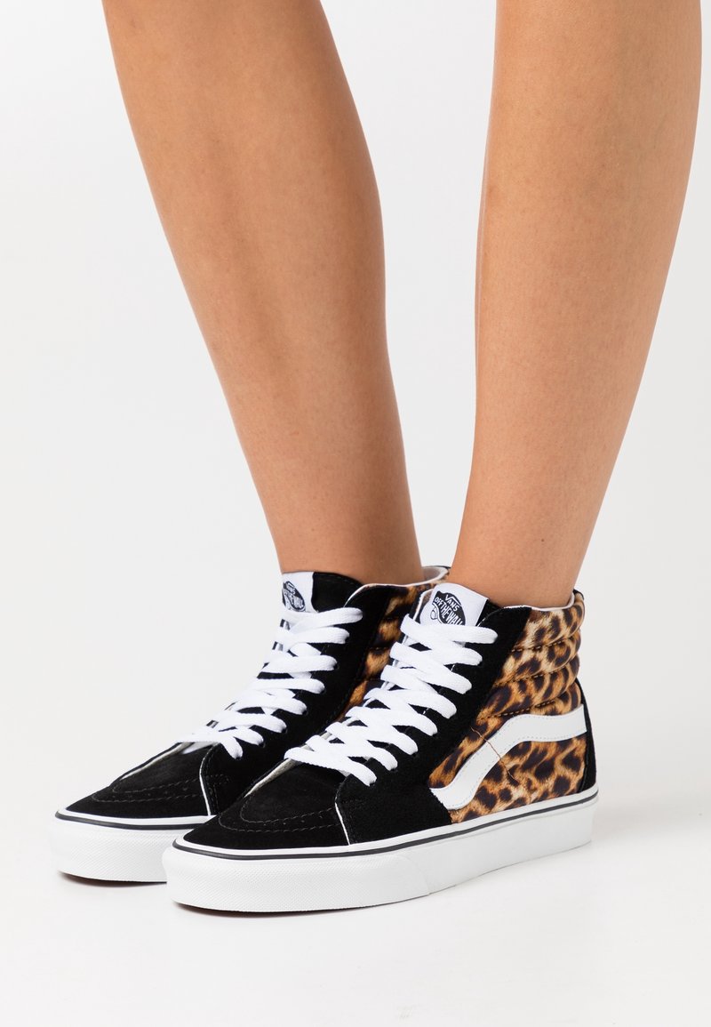 Personne portant des baskets montantes noires avec des lacets et des semelles blanches, côtés imprimé léopard, debout sur une surface blanche.