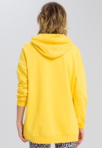 Sweat à capuche jaune en coton avec manches longues et grande capuche. Présente une coupe décontractée et des poignets et un ourlet côtelés. Le fond est uni.
