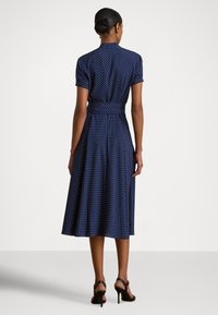 Kvinne stående med ryggen til, iført en midikjole i marineblå med hvite polkadotter, korte ermer, bred midje og svarte høye hæler.