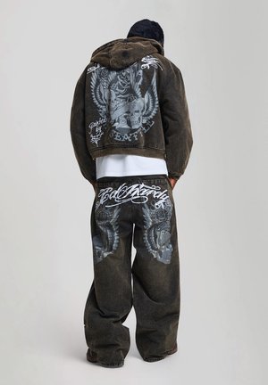 Personne portant un jean ample foncé et une veste courte à capuche assortie avec de grands motifs crâne et ailes dans le dos et sur le pantalon.