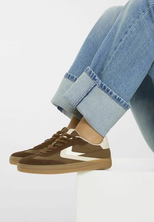 Personne portant un jean bleu retroussé et des baskets marron avec un détail en chevron blanc, assise les pieds reposant sur une surface blanche.