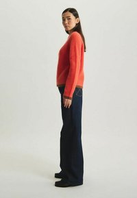 Pull en mélange de laine rouge avec texture côtelée et ourlet marron, associé à un jean à pattes d'éléphant bleu marine et des chaussures noires, vu de côté.