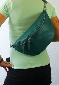 Teal leren sling bag met een gebogen ontwerp, met een ritszak en franjes. Over de schouder gedragen, met metalen hardware.