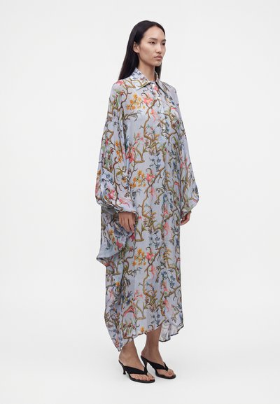 Femme portant une longue robe fluide à motifs floraux avec un ourlet asymétrique et des sandales noires à talons ouvertes, debout sur un fond blanc.