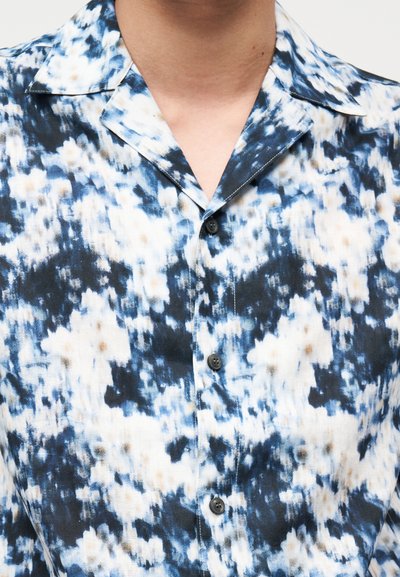 Άνδρας που φοράει μπλε και λευκό πουκάμισο με αφηρημένο μοτίβο και ανοιχτό γιακά.