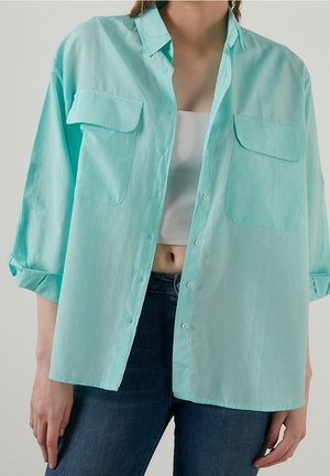 Personne portant une chemise bleu clair oversize boutonnée par-dessus un crop top blanc et un jean bleu foncé, debout devant un fond uni.