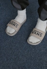 Sandales à glissière beiges avec une large sangle portant le logo "VANS", accompagnées de chaussettes blanches, sur un sol en moquette sombre.