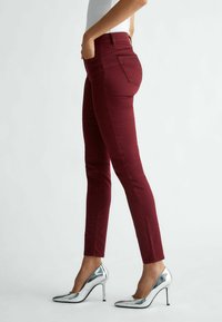 Femme portant un pantalon bordeaux slim et des chaussures à talons hauts argentées, debout les mains dans les poches devant un fond uni.