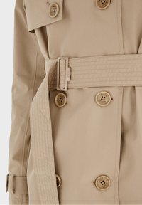 Trench-coat beige avec ceinture cousue, devant boutonné avec des boutons gravés du logo, et bracelet de manche réglable.