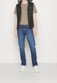 Schwarze gepolsterte Weste mit Kapuze, beiges T-Shirt, blaue Jeans und weiße Sneakers. Die Weste hat ein Logo und ist aus glattem Stoff gefertigt.