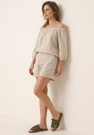 Kvinde står mod en ensfarvet væg iført en beige off-shoulder top, matchende shorts og grønne slip-on sandaler og kigger til siden.