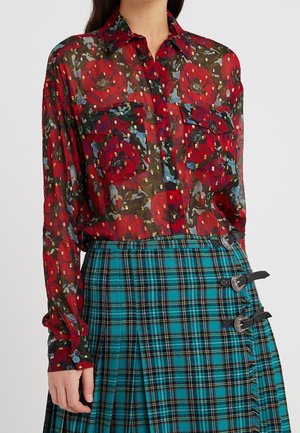 Frau trägt eine transparente rote Bluse mit Blumenmuster und langen Ärmeln sowie einen blau-grünen karierten plissierten Rock mit drei schwarzen Schnallen an der Seite.