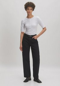 Witte top met korte mouwen en knoopdetails, gecombineerd met hooggesneden zwarte broek. Model draagt zwarte hoge hakken. Minimalistisch ontwerp.
