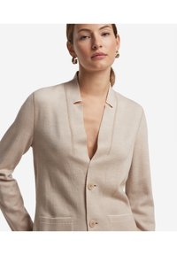 Beige kofta med notch-krage, två framfickor och träknappar. Tyget har en strukturerad yta och en avslappnad passform.