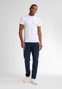 Camiseta blanca de cuello redondo con un pequeño logo en el pecho, combinada con jeans oscuros y zapatillas beige. Corte casual, diseño sencillo.